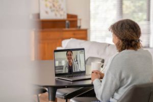 A female patent of African decent meets with her doctor remotely via a video call.  She is sitting at her kitchen table with her laptop out and a cup of coffee on the table with her.  The doctor can be seen on the screen wearing a white lab coat as she talks with her patient.